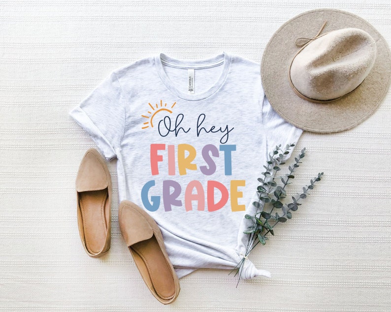 May include: A white t-shirt with the text "Oh hey first grade" in rainbow colors. The shirt is laid out on a white textured surface with a brown hat and brown shoes.