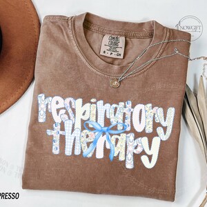 Pu&ograve; includere: T-shirt color espresso con la scritta "respiratory therapy" in un design patchwork con un fiocco blu. Una collana d'argento con un piccolo ciondolo &egrave; drappeggiata sulla maglietta. La maglietta &egrave; del marchio Comfort Colors.