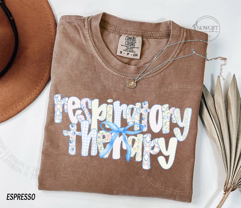 Pu&ograve; includere: T-shirt color espresso con la scritta "respiratory therapy" in un design patchwork con un fiocco blu. Una collana d'argento con un piccolo ciondolo &egrave; drappeggiata sulla maglietta. La maglietta &egrave; del marchio Comfort Colors.