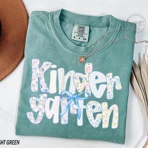 Pu&ograve; includere: T-shirt verde chiaro con la scritta "Kindergarten" in un design patchwork. Le lettere sono decorate con motivi floreali, quadretti e un fiocco blu. Una collana d'oro con un ciondolo &egrave; appoggiata sulla maglietta.