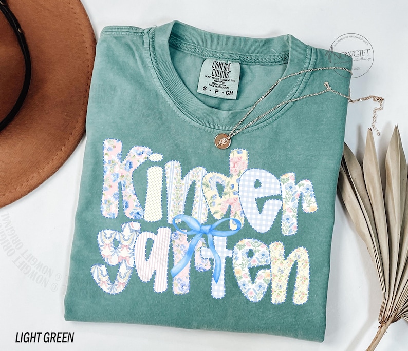 Pu&ograve; includere: T-shirt verde chiaro con la scritta "Kindergarten" in un design patchwork. Le lettere sono decorate con motivi floreali, quadretti e un fiocco blu. Una collana d'oro con un ciondolo &egrave; appoggiata sulla maglietta.