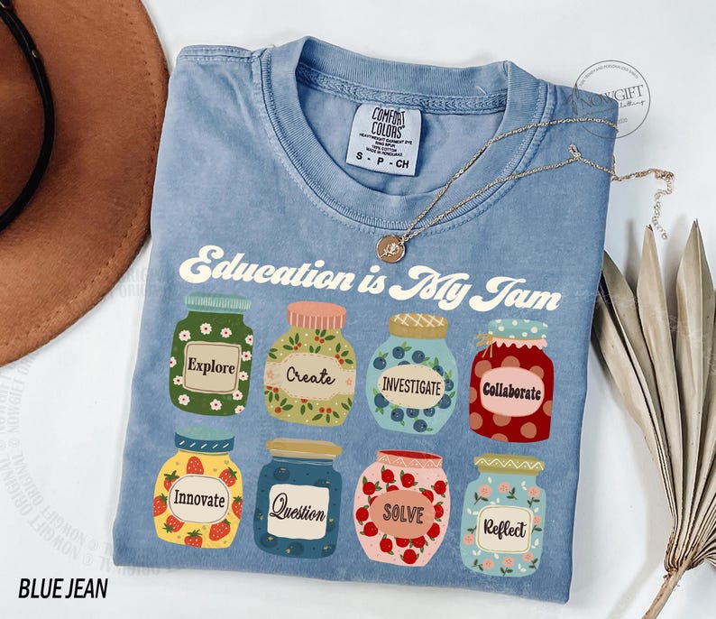 May include: A blue jean t-shirt with the text "Education is My Jam" printed on the front. The text is surrounded by illustrations of jars with different words on them, such as "Explore", "Create", "Investigate", "Collaborate", "Innovate", "Question", "Solve", and "Reflect".