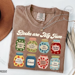 May include: A brown t-shirt with the slogan "Books are My Jam" and illustrated jam jars, each with a book-related label. The shirt has a crew neck and short sleeves. A silver necklace is draped across the shirt.