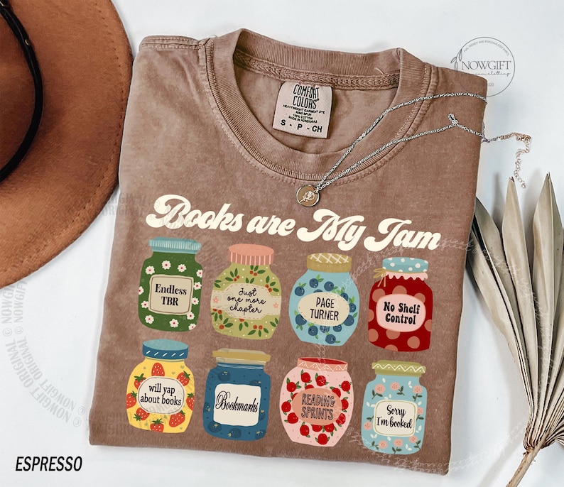 May include: A brown t-shirt with the slogan "Books are My Jam" and illustrated jam jars, each with a book-related label. The shirt has a crew neck and short sleeves. A silver necklace is draped across the shirt.