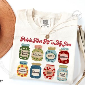 May include: Ivory-colored t-shirt with the text "Pelvic Floor PT is My Jam" in red script. The shirt features illustrations of jars with labels like "Perinatal Health" and "Kegels". A gold necklace is draped on the shirt.