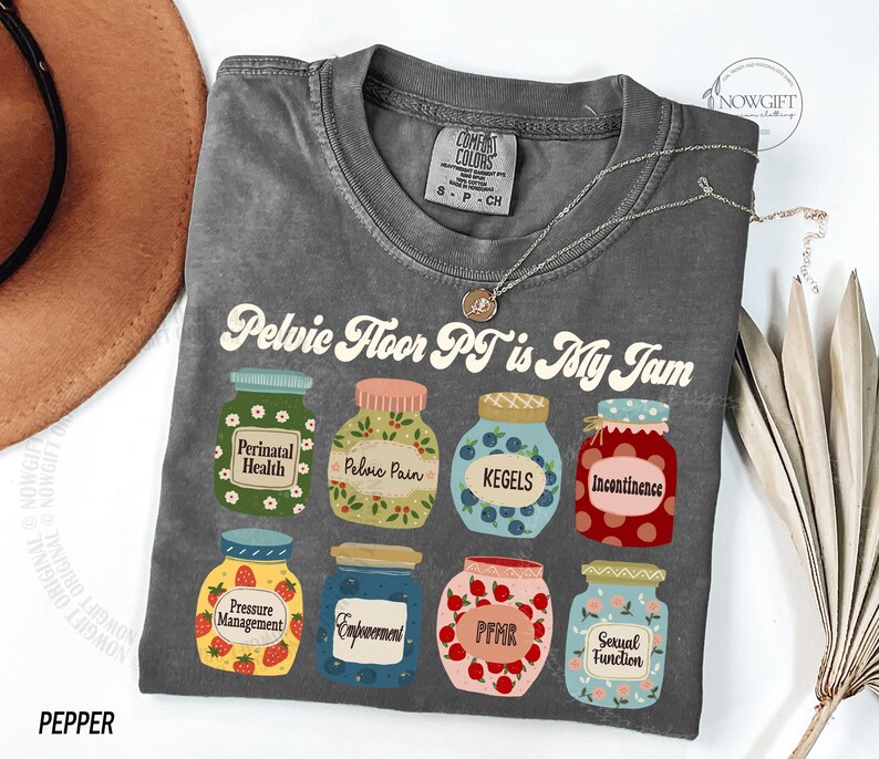 May include: A gray t-shirt with the text "Pelvic Floor PT is My Jam" and illustrations of jam jars. Each jar has a different label, including "Perinatal Health," "Pelvic Pain," and "Kegels." A silver necklace is draped across the shirt.