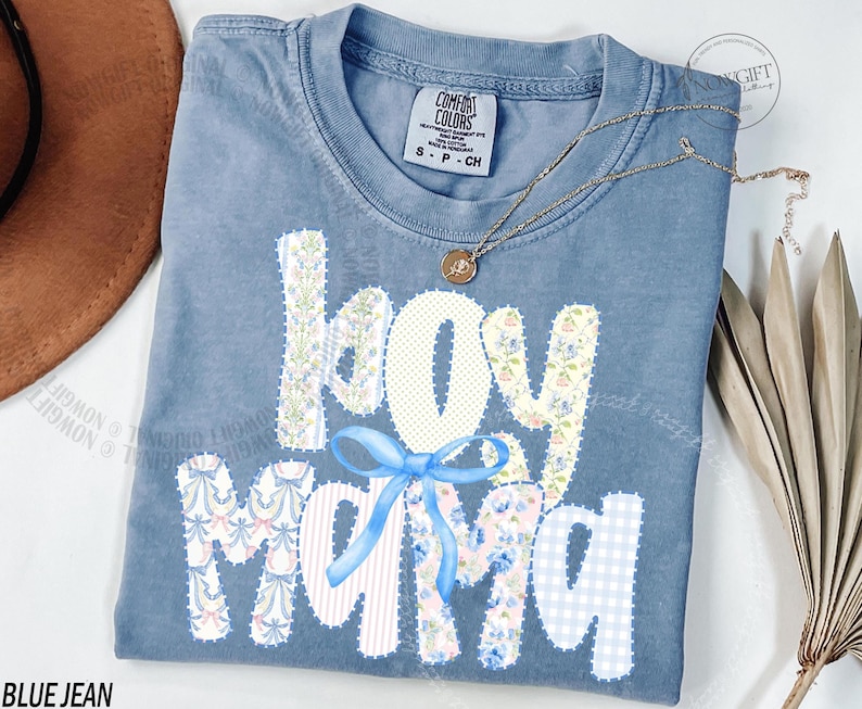 Puede incluir: Camiseta azul vaquero Comfort Colors con las palabras "boy mama" en un dise&ntilde;o de patchwork. Las letras est&aacute;n decoradas con patrones florales, a rayas y de cuadros, y un lazo azul. Un collar dorado con un colgante est&aacute; drapeado sobre la camisa.