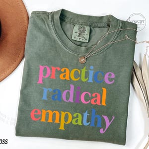May include: Olive green t-shirt with the words "practice radical empathy" in colorful letters. A gold necklace with a pendant rests on the shirt. The shirt is labeled "MOSS" at the bottom left.