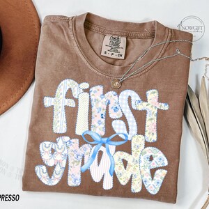 May include: Espresso-colored t-shirt with the words "first grade" in a patchwork design with floral and gingham patterns. A light blue bow is below the words. A silver necklace with a pendant is on the shirt.