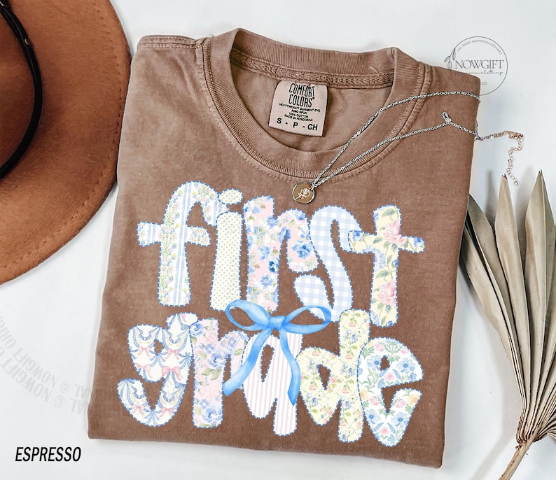 May include: Espresso-colored t-shirt with the words "first grade" in a patchwork design with floral and gingham patterns. A light blue bow is below the words. A silver necklace with a pendant is on the shirt.