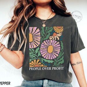 May include: A dark gray t-shirt with a floral design in pink, yellow, and green. The text "PEOPLE OVER PROFIT" is printed on the shirt.