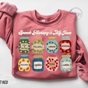 May include: A pink sweatshirt with the text "Speech Therapy is My Jam" in a retro font. The text is surrounded by illustrations of jars of jam, each representing a different aspect of speech therapy, such as voice and hearing, articulation, language, stuttering, swallowing, cognition, social, and modalities.
