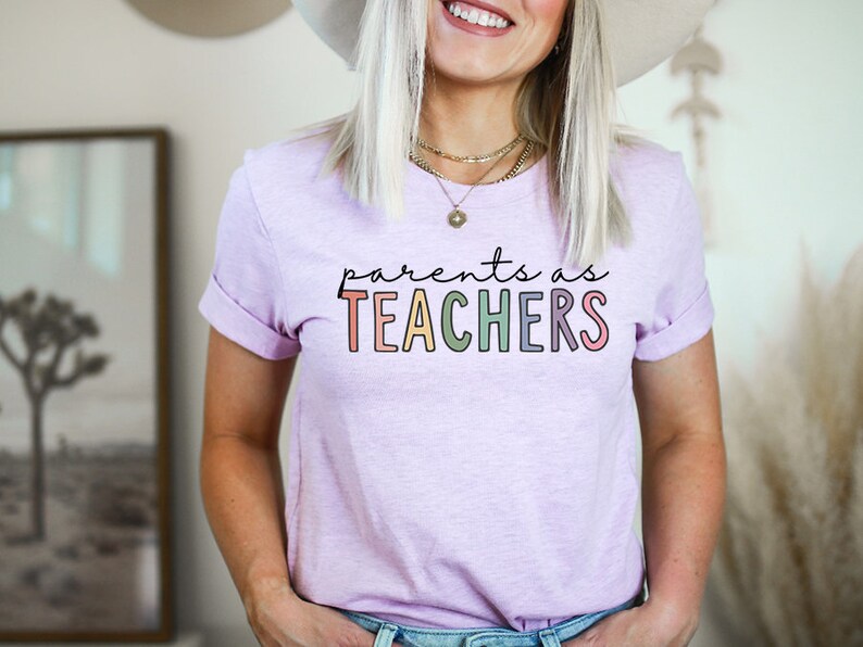 Puede incluir: Una camiseta de color morado claro con el texto "parents as TEACHERS" en colores arco&iacute;ris.