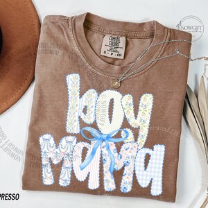 Puede incluir: Camiseta color espresso con las palabras "boy mama" en un dise&ntilde;o de patchwork. Las letras est&aacute;n adornadas con patrones florales y de cuadros, y un lazo azul claro. Un collar plateado con un colgante descansa sobre la camisa.