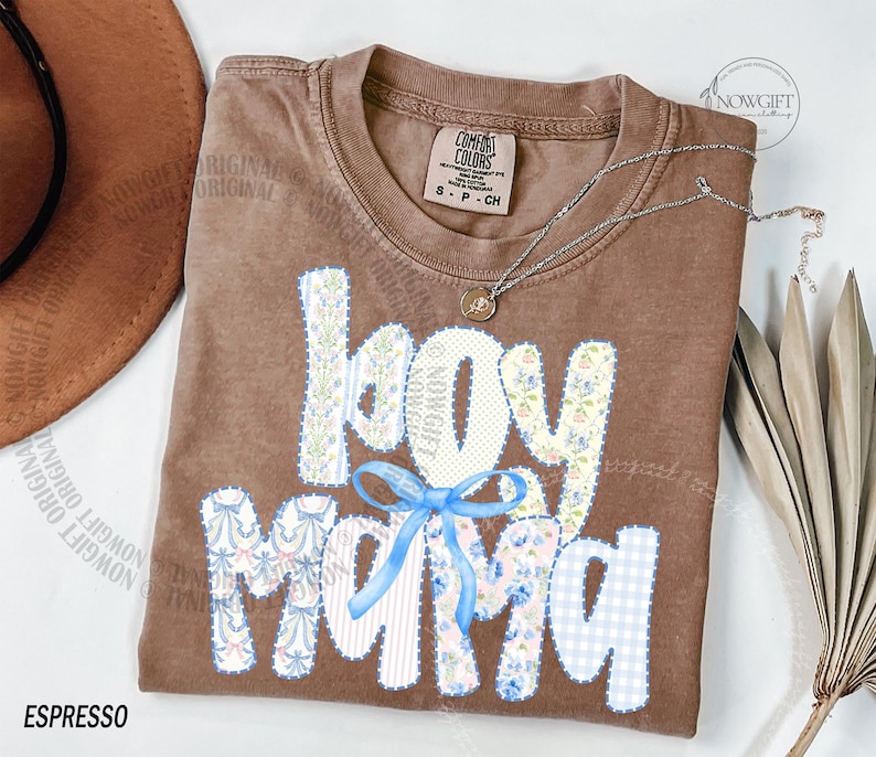 Puede incluir: Camiseta color espresso con las palabras "boy mama" en un dise&ntilde;o de patchwork. Las letras est&aacute;n adornadas con patrones florales y de cuadros, y un lazo azul claro. Un collar plateado con un colgante descansa sobre la camisa.