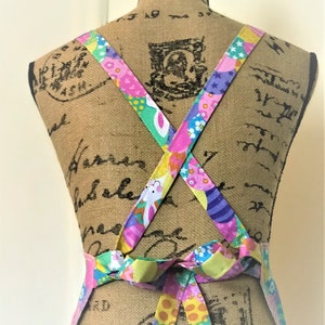 May include: A colorful Easter-themed apron with a criss-cross back tie. The apron features a pattern of pastel-colored eggs, bunnies, and polka dots.