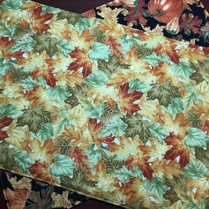 May include: A set of two fall-themed placemats featuring a pattern of colorful autumn leaves in shades of green, yellow, orange, and brown. The placemats are made of fabric and have a white background.