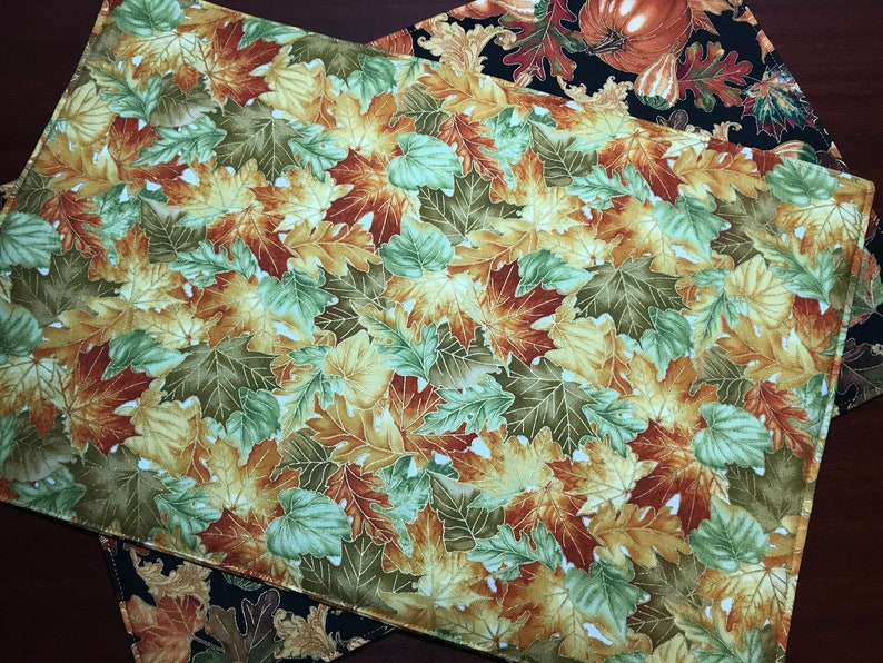 May include: A set of two fall-themed placemats featuring a pattern of colorful autumn leaves in shades of green, yellow, orange, and brown. The placemats are made of fabric and have a white background.
