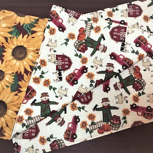 May include: Two fabric placemats with a fall theme. One placemat features a red background with yellow sunflowers. The other placemat has a white background with red barns, scarecrows, and trucks carrying pumpkins.