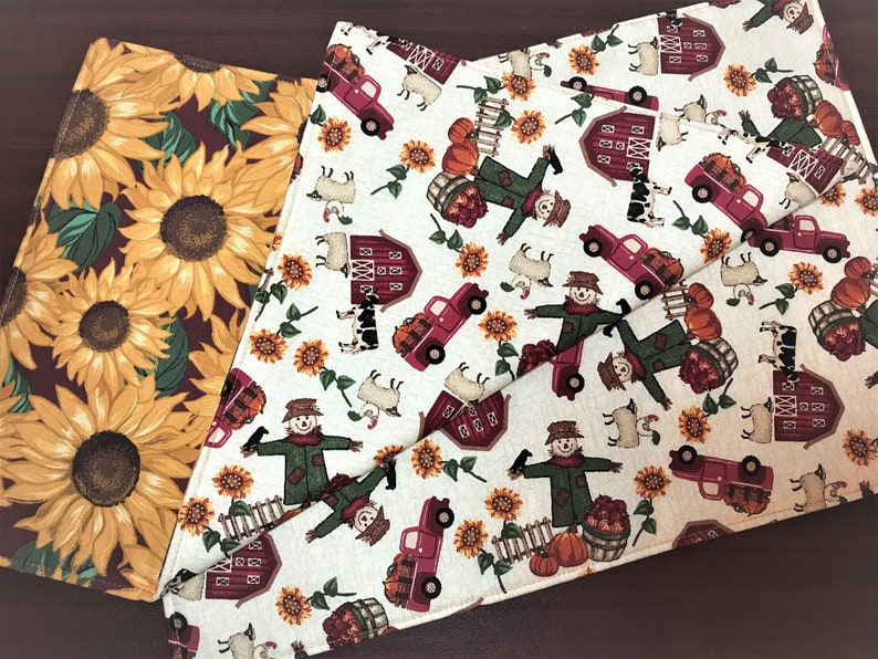 May include: Two fabric placemats with a fall theme. One placemat features a red background with yellow sunflowers. The other placemat has a white background with red barns, scarecrows, and trucks carrying pumpkins.