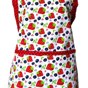 May include: A white apron with a red and white polka dot trim. The apron has a pocket and is decorated with a pattern of strawberries and blueberries.