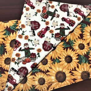 May include: Two fabric placemats with a fall theme. One placemat features a white background with red barns, scarecrows, pumpkins, and trucks. The other placemat features a burgundy background with yellow sunflowers.