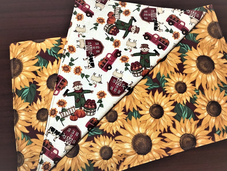 May include: Two fabric placemats with a fall theme. One placemat features a white background with red barns, scarecrows, pumpkins, and trucks. The other placemat features a burgundy background with yellow sunflowers.