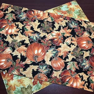 May include: Two black fabric placemats with a gold and brown autumn leaf and pumpkin pattern. The placemats are made of a soft, quilted fabric.