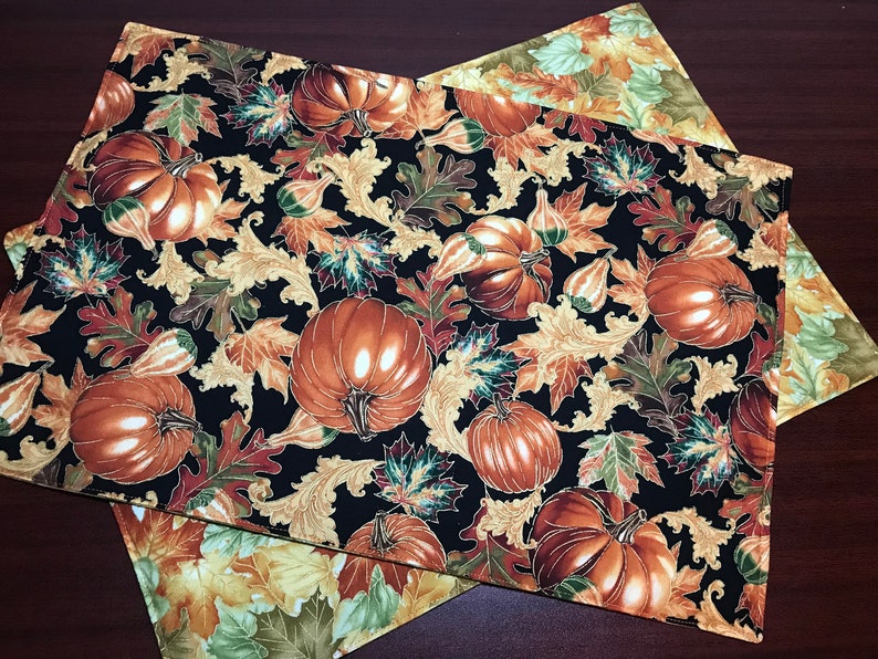 May include: Two black fabric placemats with a gold and brown autumn leaf and pumpkin pattern. The placemats are made of a soft, quilted fabric.