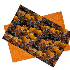 May include: Two orange and white fabric placemats with a black background featuring a pattern of pumpkins and fall foliage.