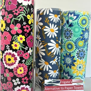 May include: Three cylindrical rolls of reusable fabric towels featuring distinct patterns. The left roll has a black background with pink, yellow, and white flowers. The center roll is navy blue with white daisies. The right roll has a light green background with blue and yellow floral mandalas. A sign reads Frigate's Alernative to Paper Towels.