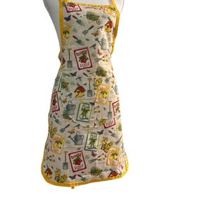 May include: A cream-colored apron with a vibrant garden-themed print featuring illustrations of seed packets, watering cans, shovels, butterflies, and birdhouses. The apron includes a front pocket and is finished with bright yellow trim.