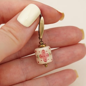 Peut inclure: Boucle d'oreille unique avec une perle cylindrique crème ornée d'un motif floral rose. La boucle d'oreille a un dessus en métal doré et une petite perle ronde dorée en bas. La boucle d'oreille est tenue dans une main avec du vernis à ongles jaune pâle.
