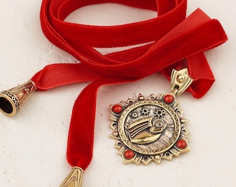 May include: A red velvet ribbon necklace with gold-toned metal accents. The necklace features a circular pendant with a bird design and red gemstone embellishments. The ribbon ends have decorative metal caps.