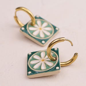 May include: Gold-coloured hoop earrings with square ceramic pendants. Each pendant is teal with a white daisy design. The earrings are on a light-coloured surface.