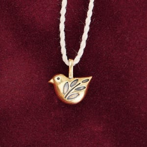 Può includere: Un ciondolo a forma di uccello dorato con un cordino bianco. L'uccello presenta un design dettagliato con motivi a forma di foglia sull'ala. Il ciondolo è sospeso su uno sfondo rosso scuro.