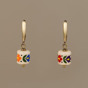 May include: A pair of gold-tone earrings with a white, cylindrical bead. Each bead is decorated with a colorful floral pattern in orange, blue, and red. The earrings have a lever-back closure.
