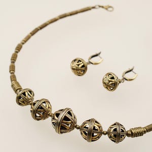 May include: A gold-toned necklace and matching earrings set. The necklace features large, openwork spherical beads interspersed with smaller beads. The earrings have a similar design. The jewelry is displayed against a light background.