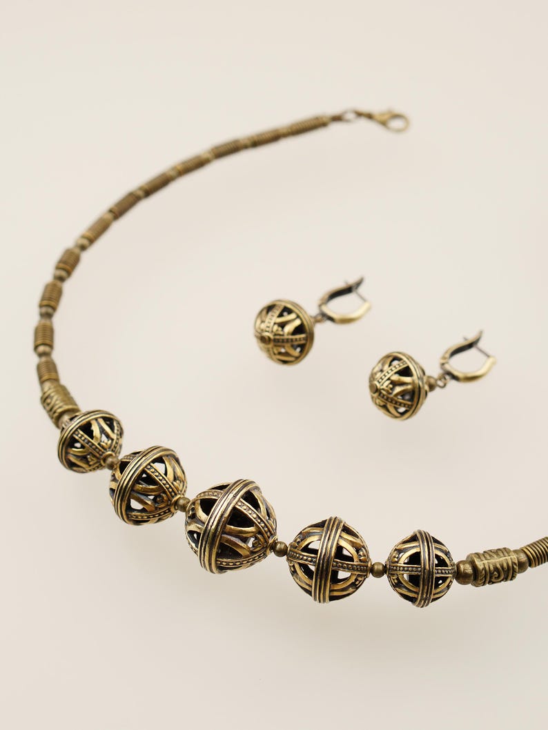 May include: A gold-toned necklace and matching earrings set. The necklace features large, openwork spherical beads interspersed with smaller beads. The earrings have a similar design. The jewelry is displayed against a light background.
