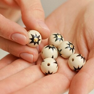 Hands holding small hand-painted ceramic beads with an earthy white base, black star-like folk motifs, and fine gold details, showing the handmade bead-making process against a soft neutral background.