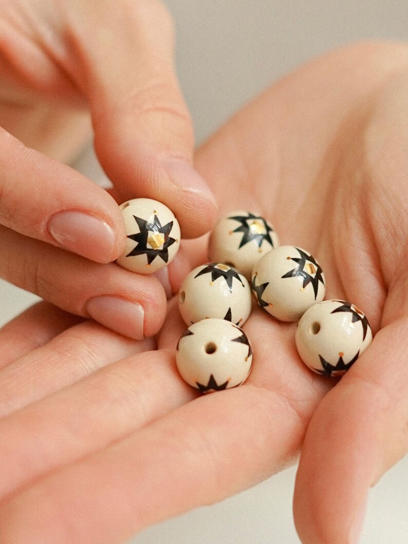 Hands holding small hand-painted ceramic beads with an earthy white base, black star-like folk motifs, and fine gold details, showing the handmade bead-making process against a soft neutral background.