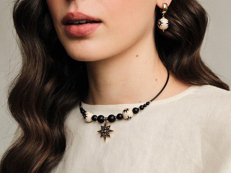 May include: A black and white beaded choker necklace with a gold star pendant. The necklace is made of black and white beads with a gold star pendant in the center. The star pendant has a black center and gold points.