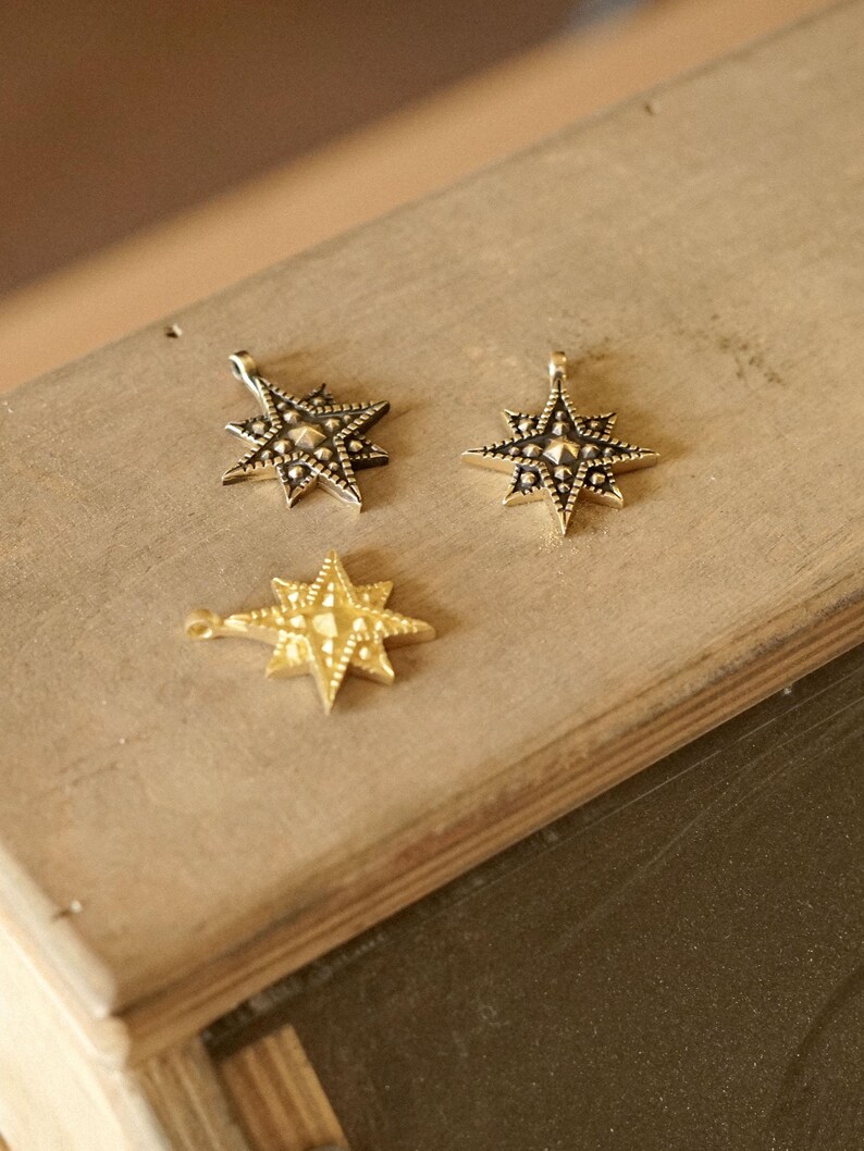 Three small star-shaped brass charms with detailed geometric engraving, shown in antique gold and darker brass tones, arranged on a wooden work surface under warm natural light.