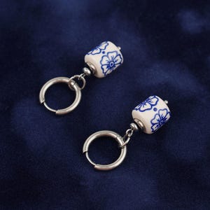 May include: A pair of earrings featuring white ceramic beads with blue floral patterns. The beads are cylindrical and hang from silver-colored hoop earrings. The background is a dark blue fabric.