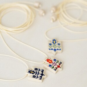 Set of three handmade ceramic Tryzub necklaces with folk floral designs in blue, red, and navy, each on a white base with cream adjustable cords and ceramic bead ends.