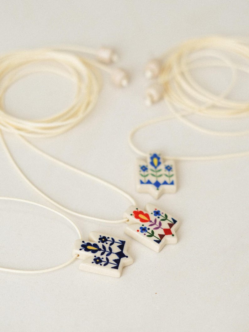 Set of three handmade ceramic Tryzub necklaces with folk floral designs in blue, red, and navy, each on a white base with cream adjustable cords and ceramic bead ends.