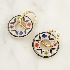 Puede incluir: Un par de pendientes de aro dorados con un colgante circular de color crema. El colgante está decorado con un borde negro, hojas negras y flores coloridas en rojo, azul y verde. Los pendientes se exhiben sobre una superficie de mármol blanco.