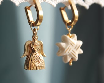 May include: A pair of gold earrings with a white ceramic star and a gold angel pendant. The angel is wearing a dress with a geometric pattern.