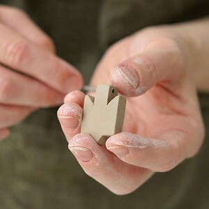 Close-up of hands refining the shape of an unglazed ceramic Tryzub pendant, showing the detailed craftsmanship involved in the handmade jewelry process.