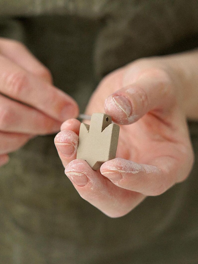Close-up of hands refining the shape of an unglazed ceramic Tryzub pendant, showing the detailed craftsmanship involved in the handmade jewelry process.